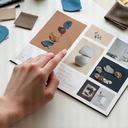 Ang kamay ng isang tagadisenyo ay nagtuturo sa isang mood board na may mga larawan ng ceramic at glass sa isang table, sumisimbolo sa creative direction.